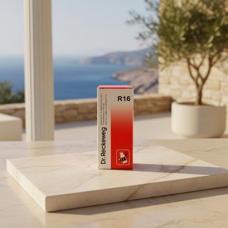 A slim rectangular Dr. Reckeweg R16 product box stands upright on a light marble tray outdoors on a sunlit table, with a blurred coastal terrace and potted tree in the background.