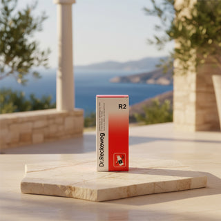 A tall rectangular product box for Dr. Reckeweg R2, featuring a white-to-red gradient design with black text and a small red-and-white icon near the bottom, set against a pale pink background.